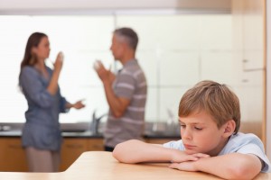 sad boy sitting at table with parents arguing in the background