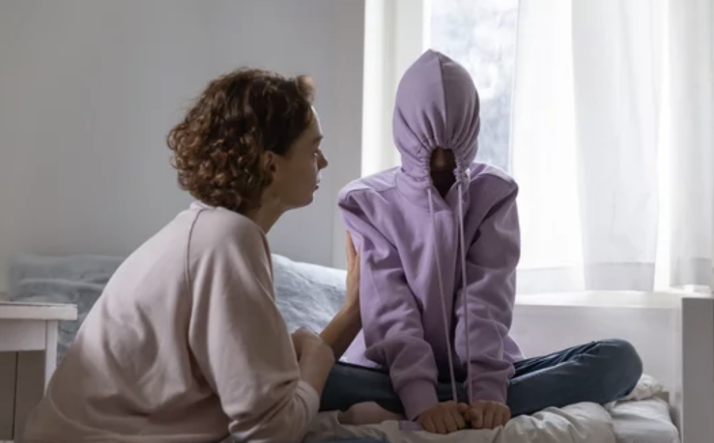 Depressed teen girl with her hoodie covering her face and mother comforting teen.