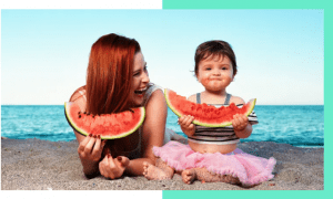 Mother and toddler eating watermelon at beach smiling and laughing.